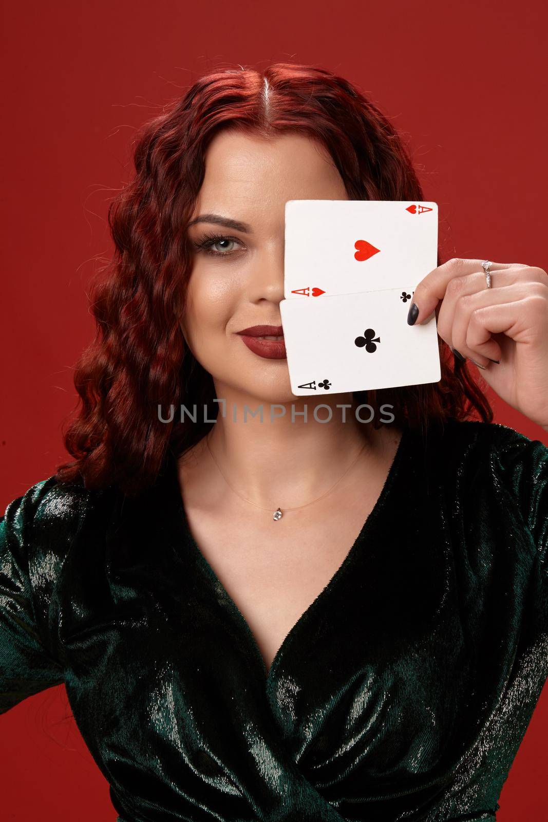 Pretty woman with a red curly hair, have brought playing cards to her face and smiling, on a red background. Poker
