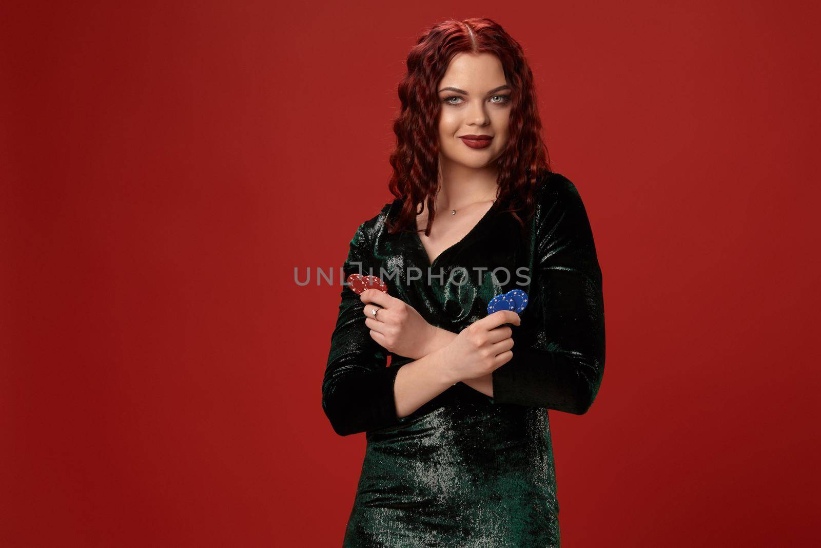 Pretty young redheaded woman is holding some gambling chips in her crossed hands and smiling, on a red background.