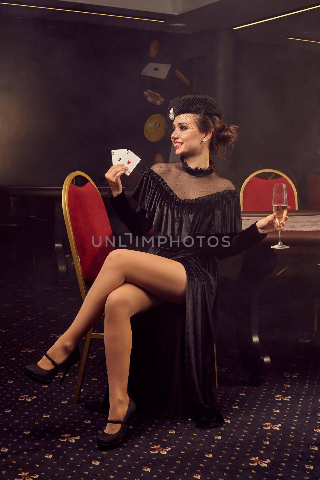 Dazzling young girl in a black velvet dress and a hat with a veil, with playing cards in her hand is looking away, smiling and posing sitting sideways on a chair against a poker table in luxury casino. Passion, cards, chips, alcohol, win, gambling - it is as female entertainment. Smoke background.