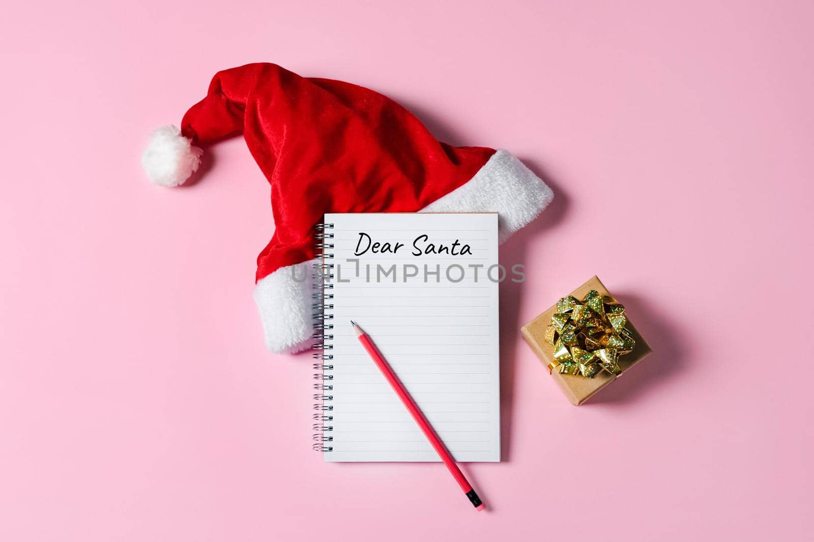 Dear Santa letter concept. Writing in Notepad letter to Santa Claus on pink background with Santa hat and gift box.