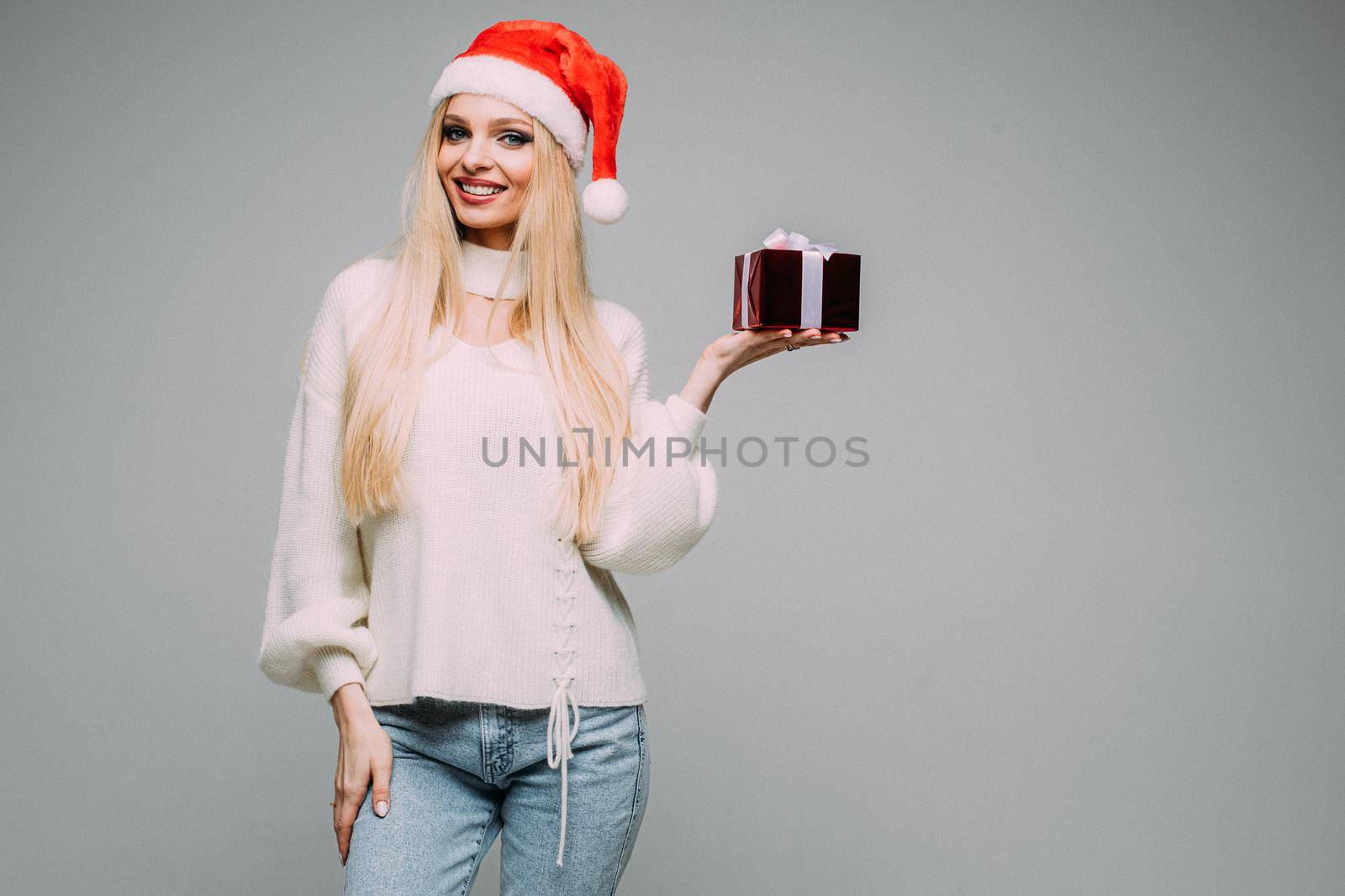 Happy young woman wearing red Santa hat and holding present box, isolated on grey background. New Year holiday concept