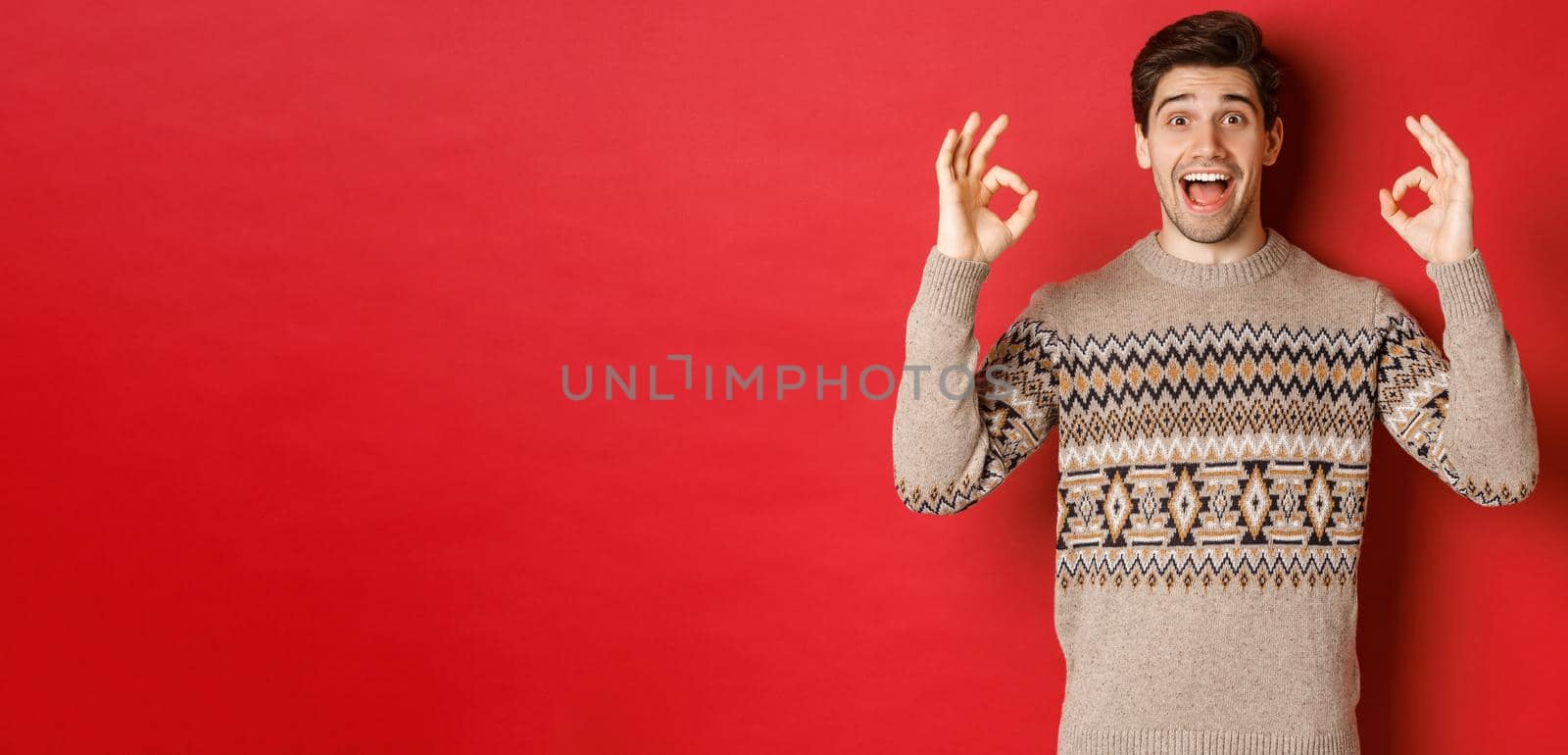 Concept of christmas celebration, winter holidays and lifestyle. Portrait of handsome man in sweater, looking amazed and showing okay signs, guarantee or recommend something good, red background.