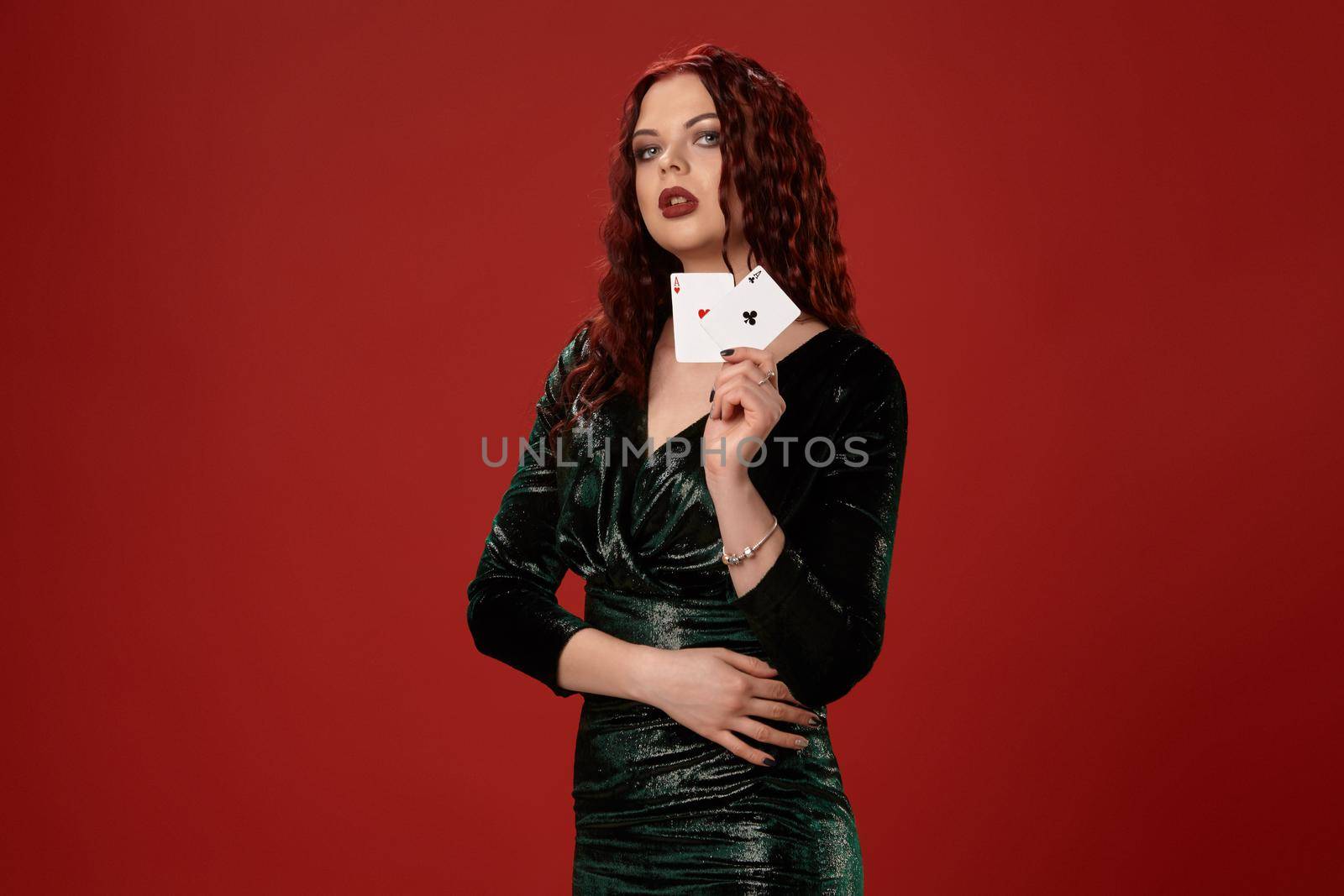 Young sexy woman with a red curly hair, holding aces in her hand and looking up, on a red background. Poker