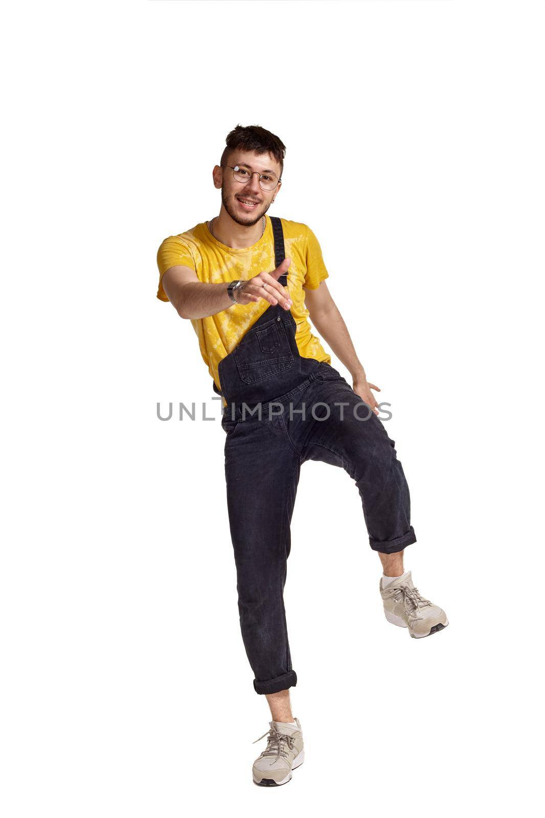 Full-length portrait of a graceful dancer in glasses, black jumpsuit, yellow t-shirt and gray sneakers fooling around in studio. Indoor photo of a man making dancing elements raising his leg isolated on white background. Music and imagination.