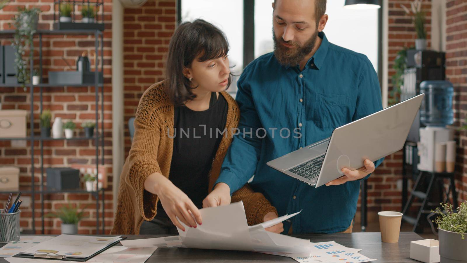 Business people checking laptop and rate charts papers to create marketing strategy. Man and woman brainstorming ideas while working on project planning and research in startup office.