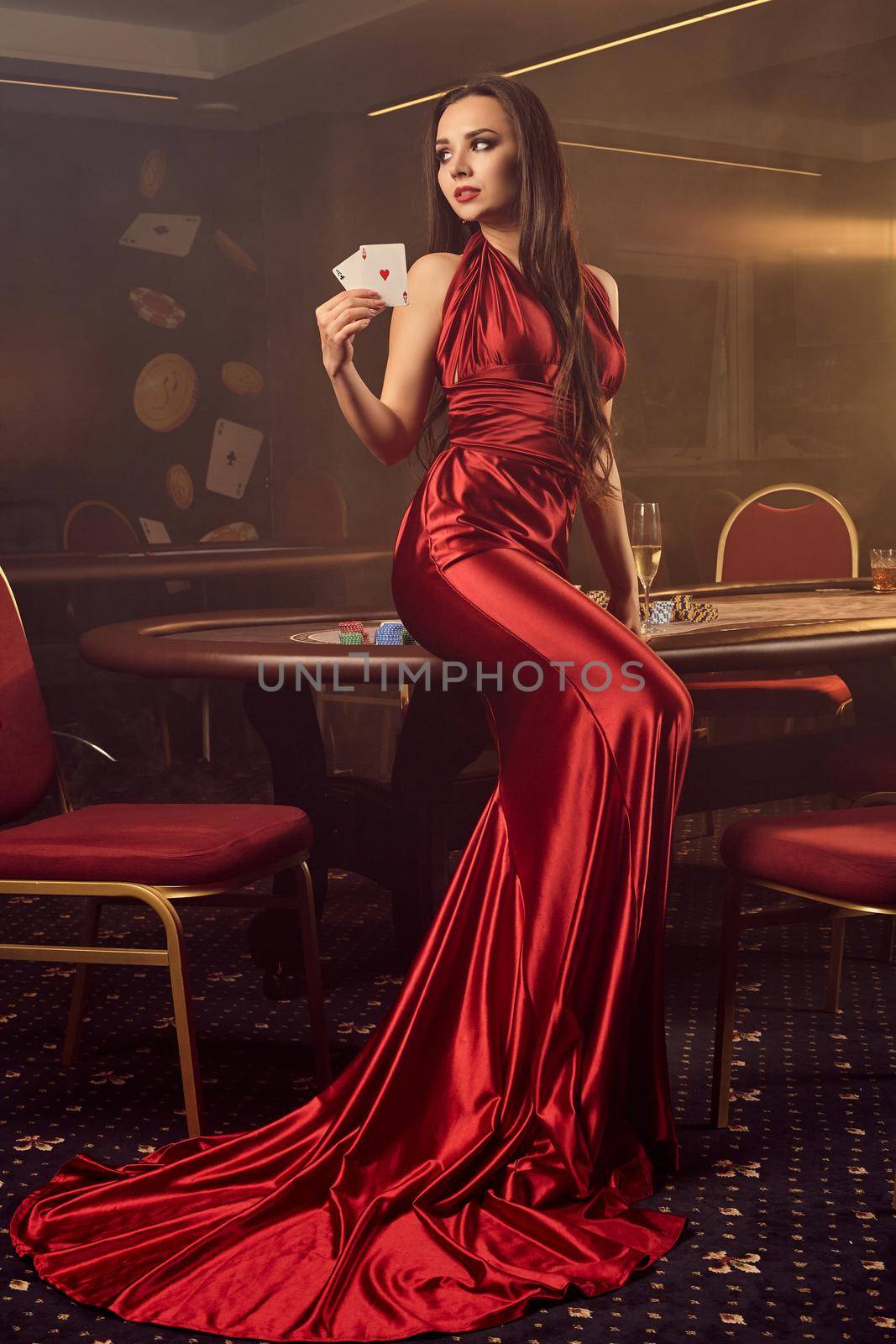 Beautiful brunette maiden in a long burgundy atlas dress, with two aces in her hand is looking back and posing sitting on a poker table in vip casino. Passion, cards, chips, alcohol drinks, win, gambling - it is a best entertainment. Smoke background.
