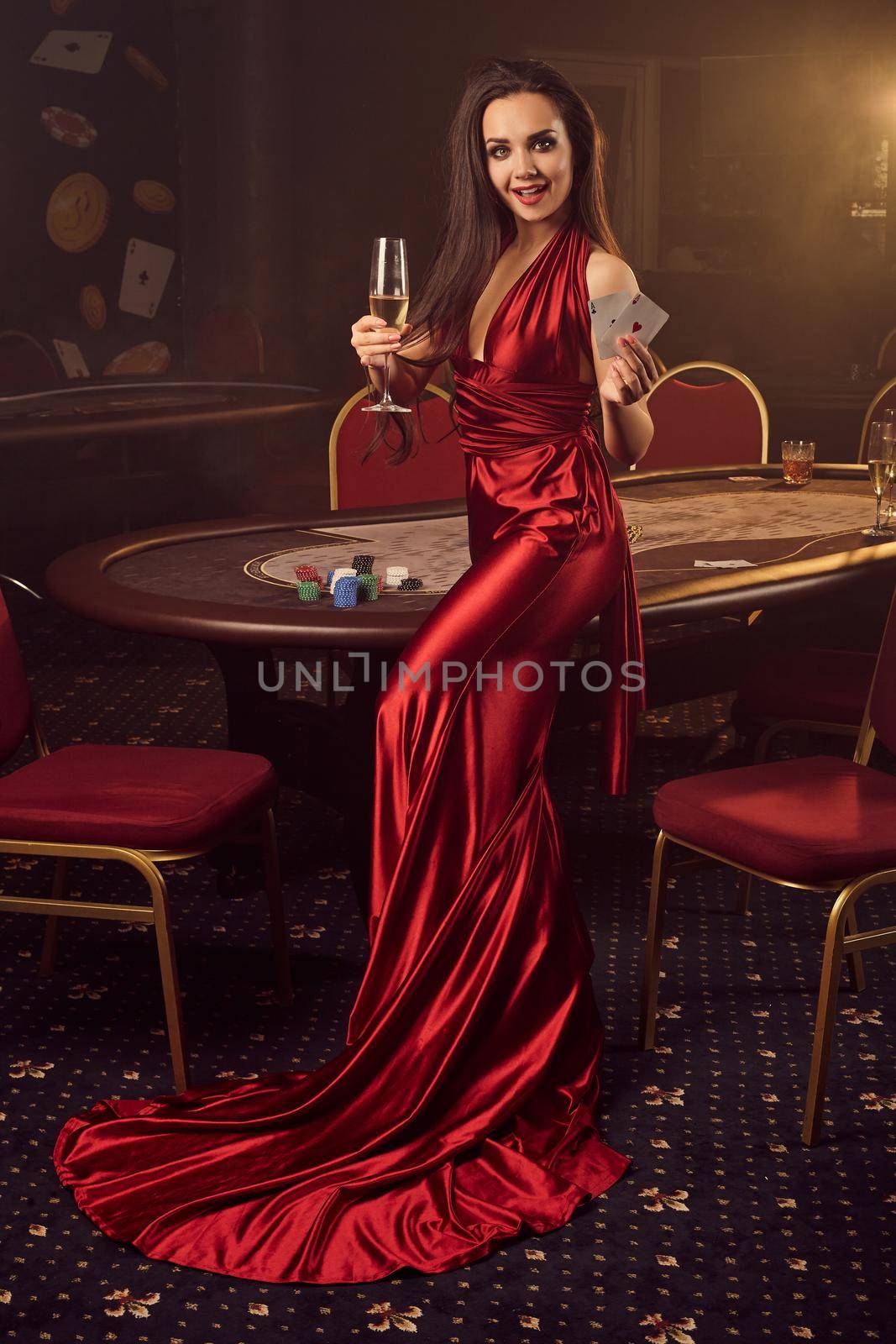 Young brunette female in a long burgundy atlas dress, with two aces and a glass of champagne in her hands is posing against a poker table in vip casino. Passion, cards, chips, alcohol drinks, win, gambling - it is a best entertainment.