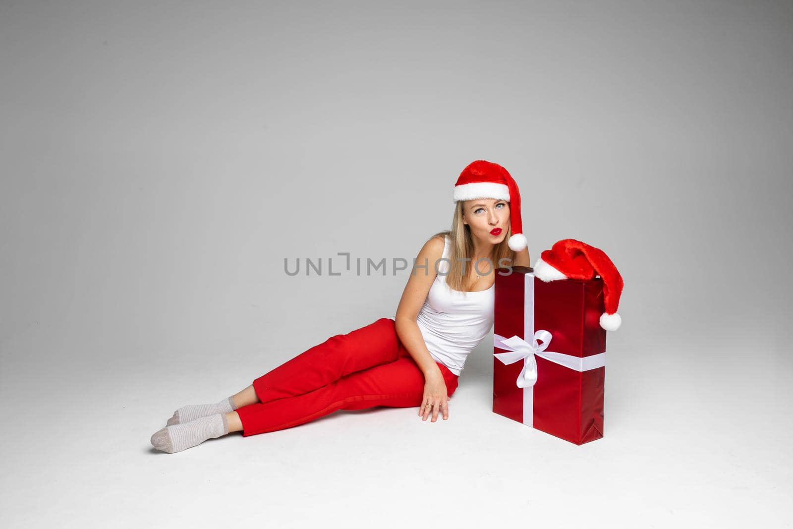 funny woman with christmas hat sits with a big red box and think what is inside, picture isolated on white background by StudioLucky