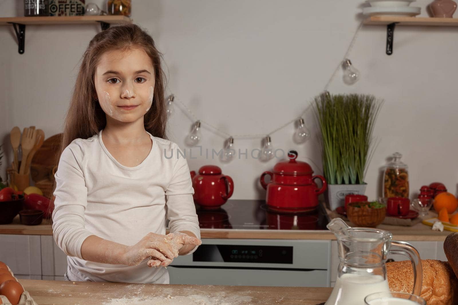 Pretty little brunette lady dressed in a white blouse is making a dough for baking bread, smiling and looking at the camera at kitchen against a white wall with shelves and bulbs on it. Happy childhood concept.
