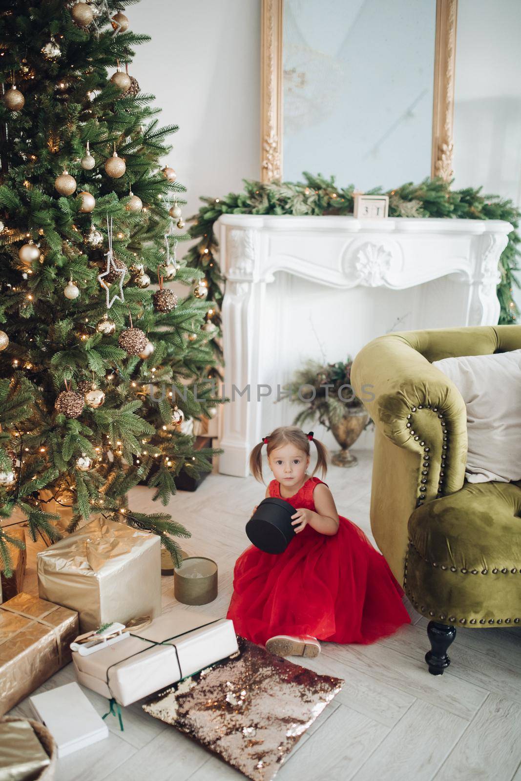 Stock photo portrait of adorable girl in red dress with festive print holding beautifully wrapped golden present in hands while sitting on the floor next to decorated Christmas treee with garland..