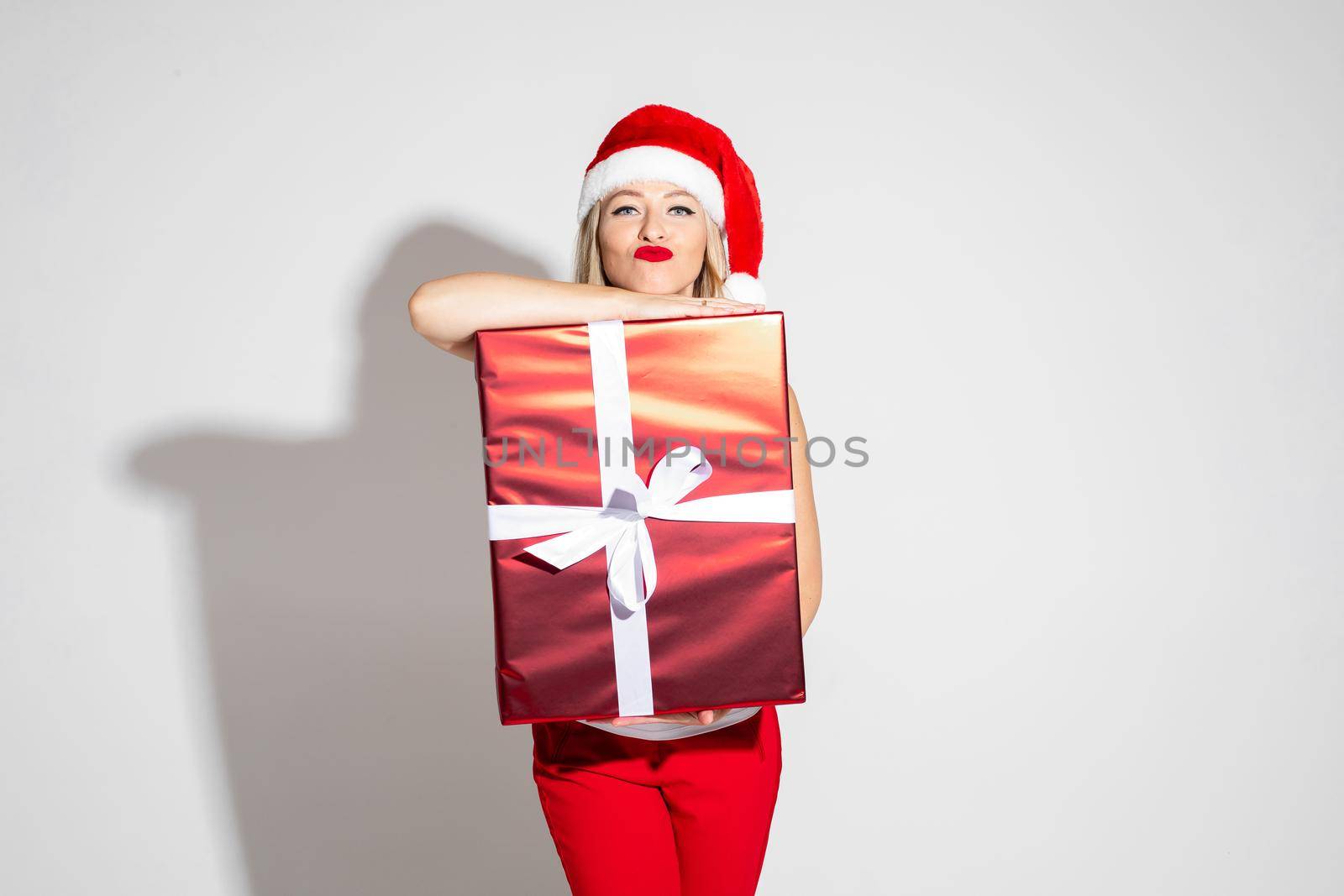 Close-up photo of a young blonde lady wearing Santa hat and red lipstic while leaning on a Christmas present. Holiday concept