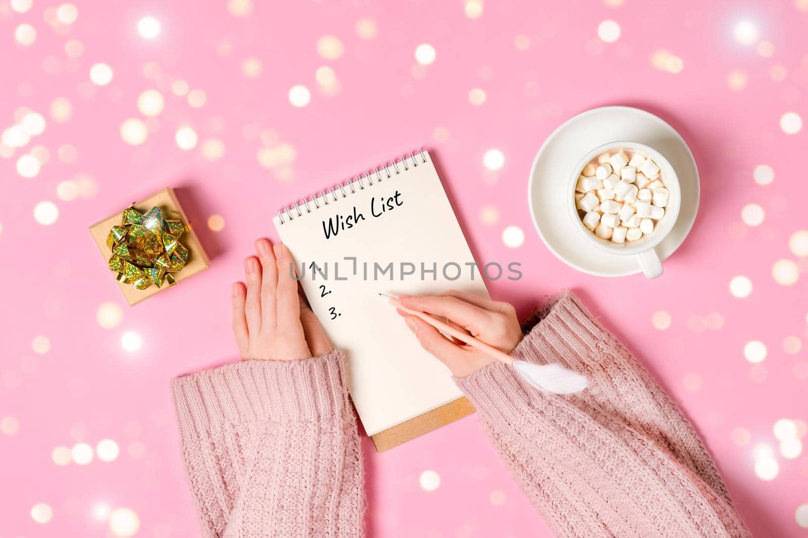 New year, Christmas or holiday wish list concept. Notepad, golden tinsel, and a cup of hot chocolate with marshmallows and female's hands writing isolated on pink background. Banner with copy space.