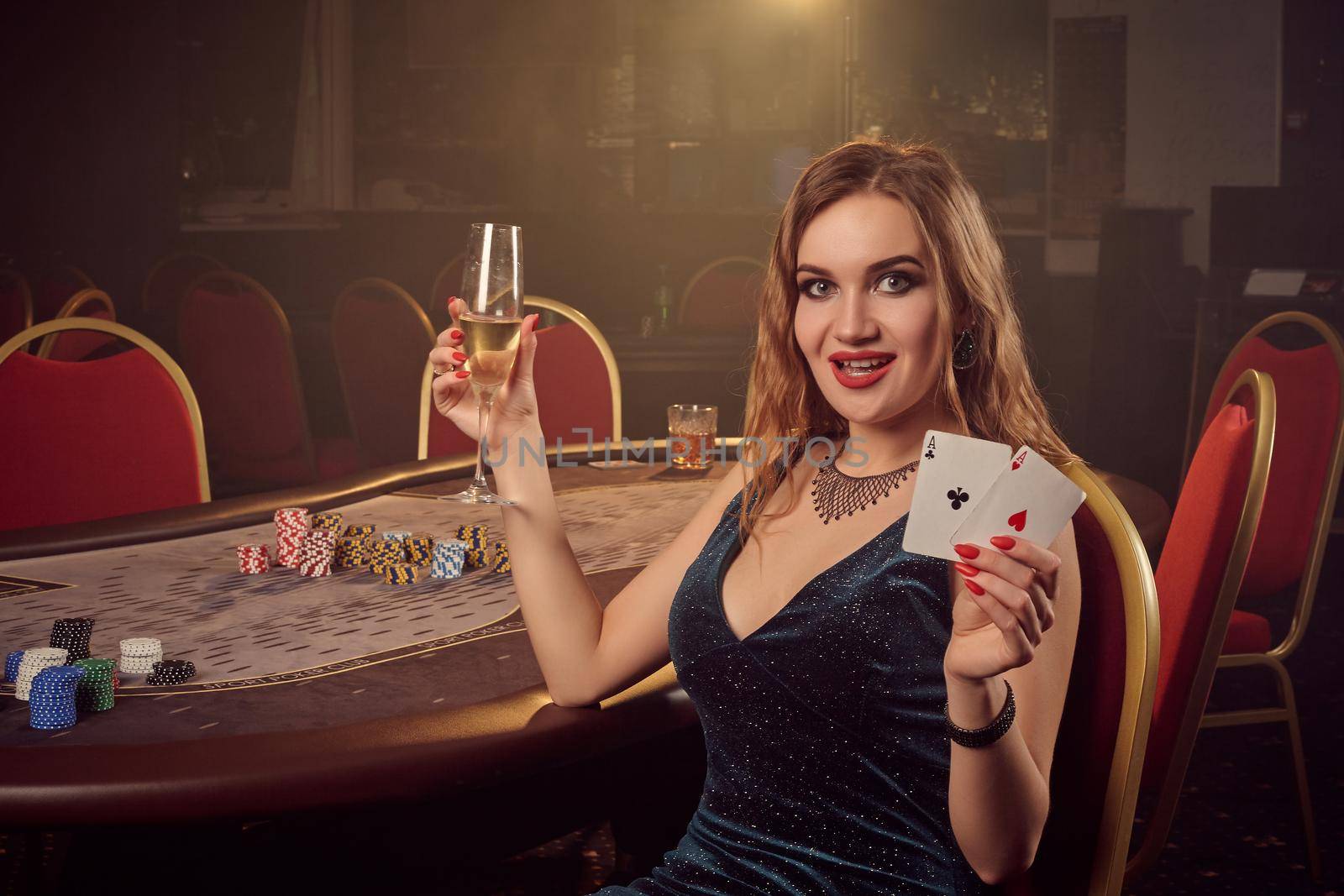 Sexy blond female in a short black dress, with two aces and a glass of champagne in her hands is posing sitting sideways on a chair against a poker table in luxury casino. Passion, cards, chips, alcohol, win, gambling - it is a female entertainment. Smoke background.