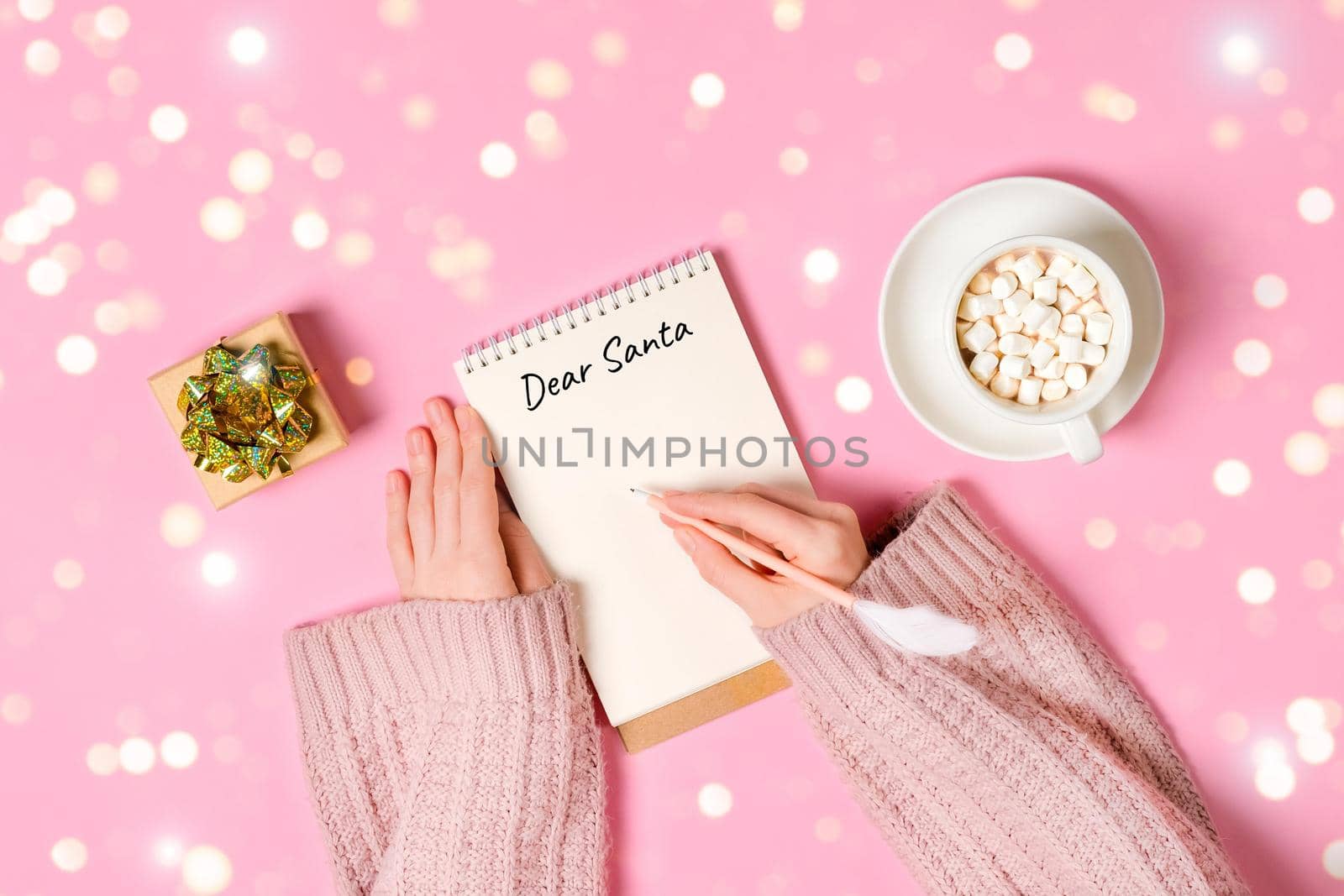 Letter to Dear Santa. Hand holding pink pen decorated with feather and writing in Notepad letter to Santa Claus on pink background with cup of hot chocolate with marshmallows, a gift, golden tinsel.