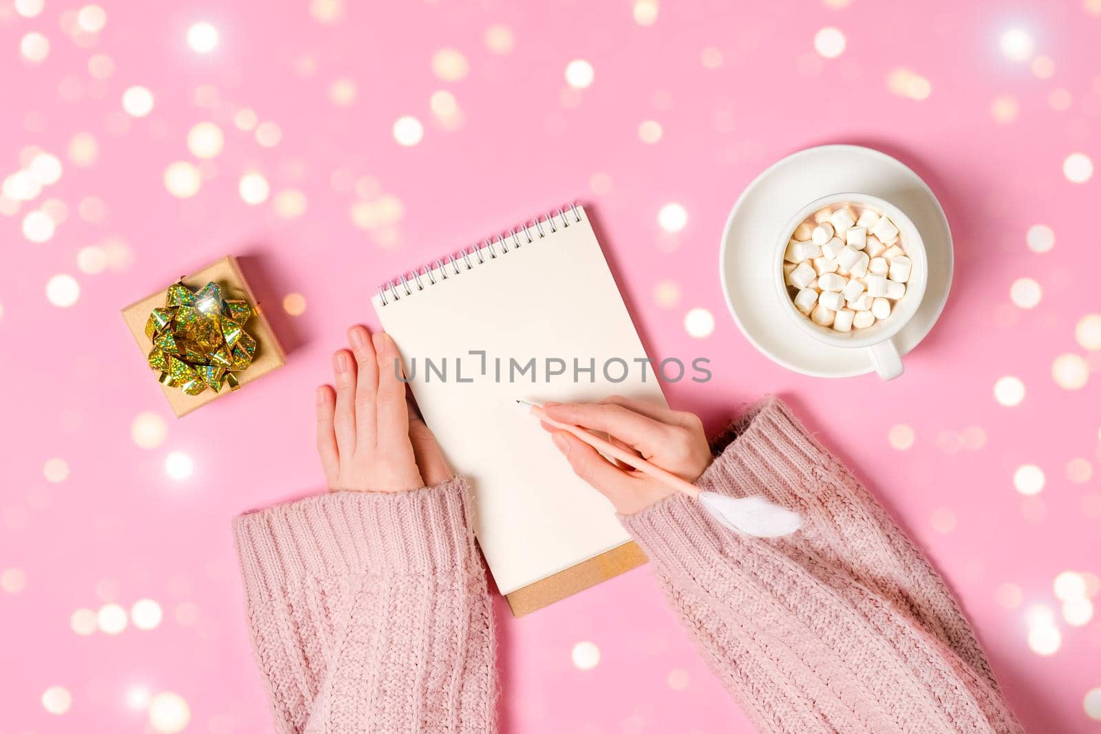 New year, Christmas or holiday wish list concept. Notepad, golden tinsel, and a cup of hot chocolate with marshmallows and female's hands writing isolated on pink background. Banner with copy space