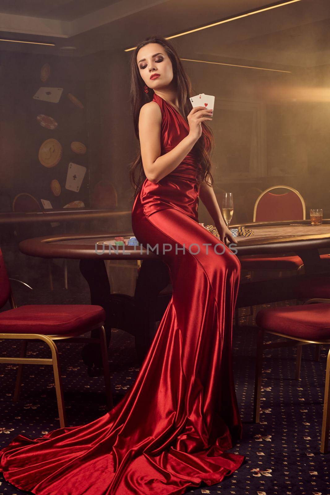 Charming brunette female in a long burgundy atlas dress, with two aces in her hand is looking down and posing sideways near a poker table in vip casino. Passion, cards, chips, alcohol drinks, win, gambling - it is a best entertainment. Smoke background.