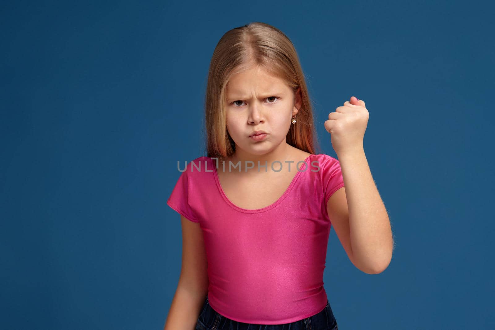 Portrait of emotional blonde little girl on blue background. in pink shirt, acting like anger unhappy person