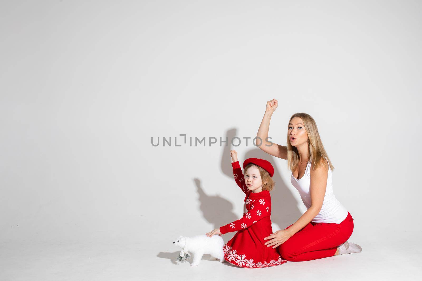 Studio photo of funny mother and daughter in red and white posing with arm up with fist imitating a train whistle. White bear toy in front of the child. Copy space.