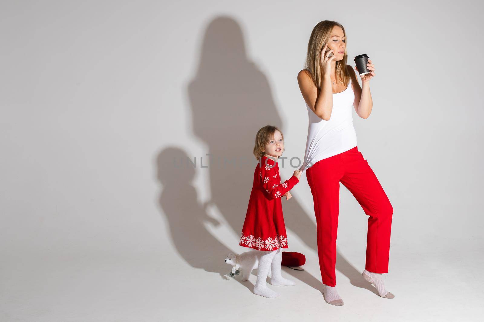 Busy mom talking via cell and drinking coffee while her daughter in red dress with Christmas pattern pulling her top demanding attention.