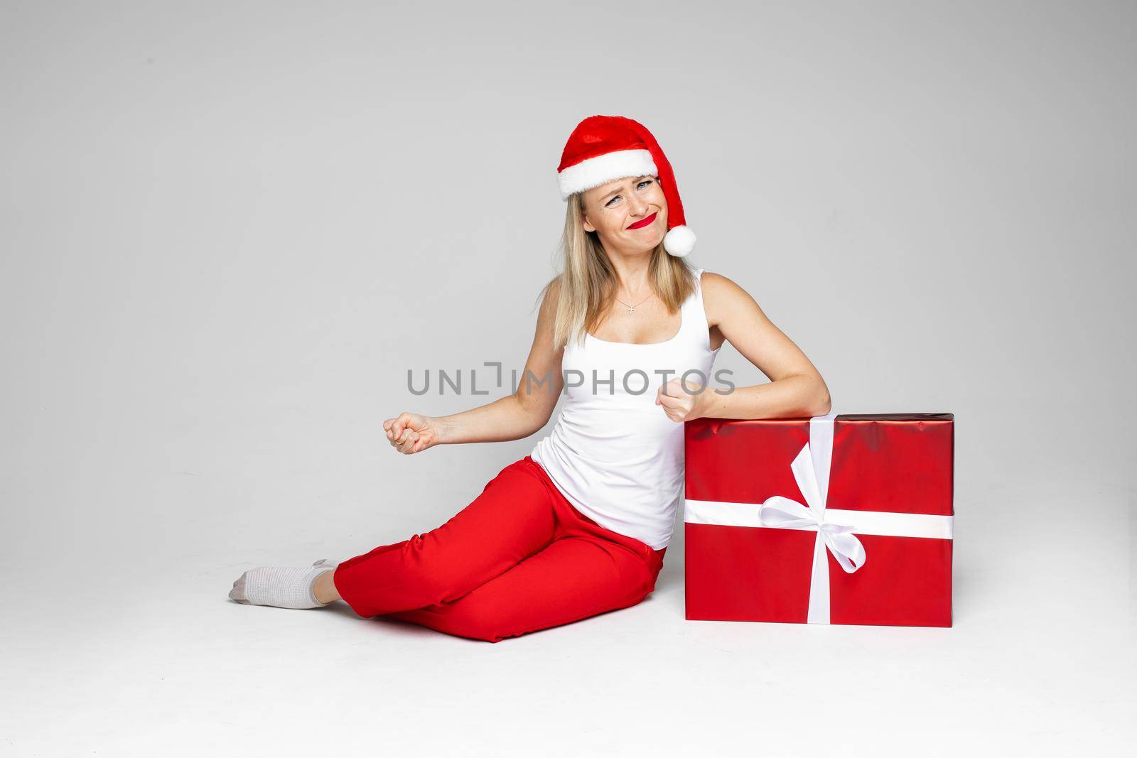 Frustrated young woman in Santa hat clenching her fists while sitting next to red boxed Christmas gift. Copy space