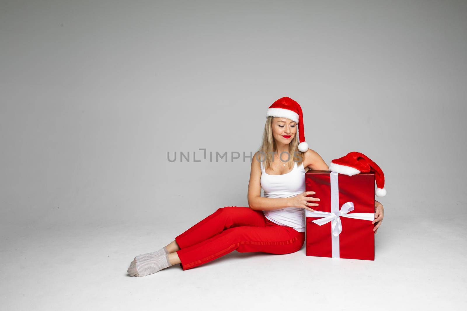 Studio photo of an amicable young lady wearing Santa hat and sitting near a nicely wrapped Christmas gift. Holiday concept