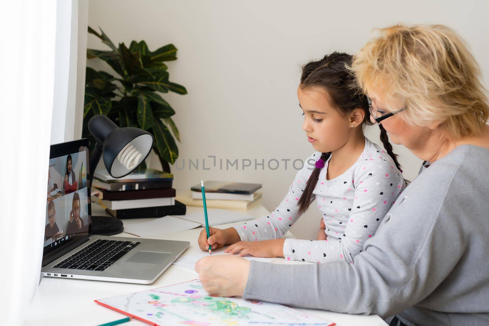 Mature grandmother helping child with homework at home. Satisfied old grandma helping her granddaughter studying in living room. Little girl writing on notebook with senior teacher sitting next to her by Andelov13