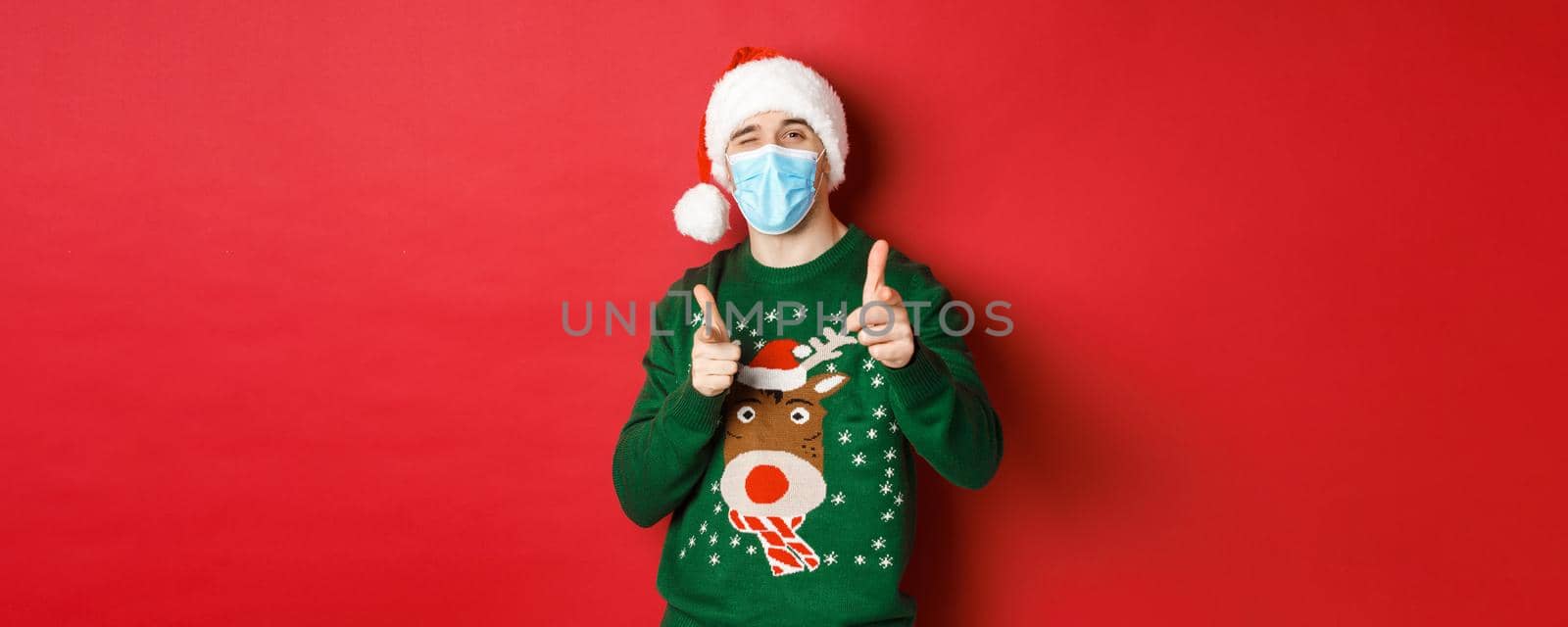 Concept of new year, covid-19 and social distancing. Cheerful man in christmas sweater, medical mask and santa hat, pointing fingers at camera, wishing happy holidays, standing over red background by Benzoix