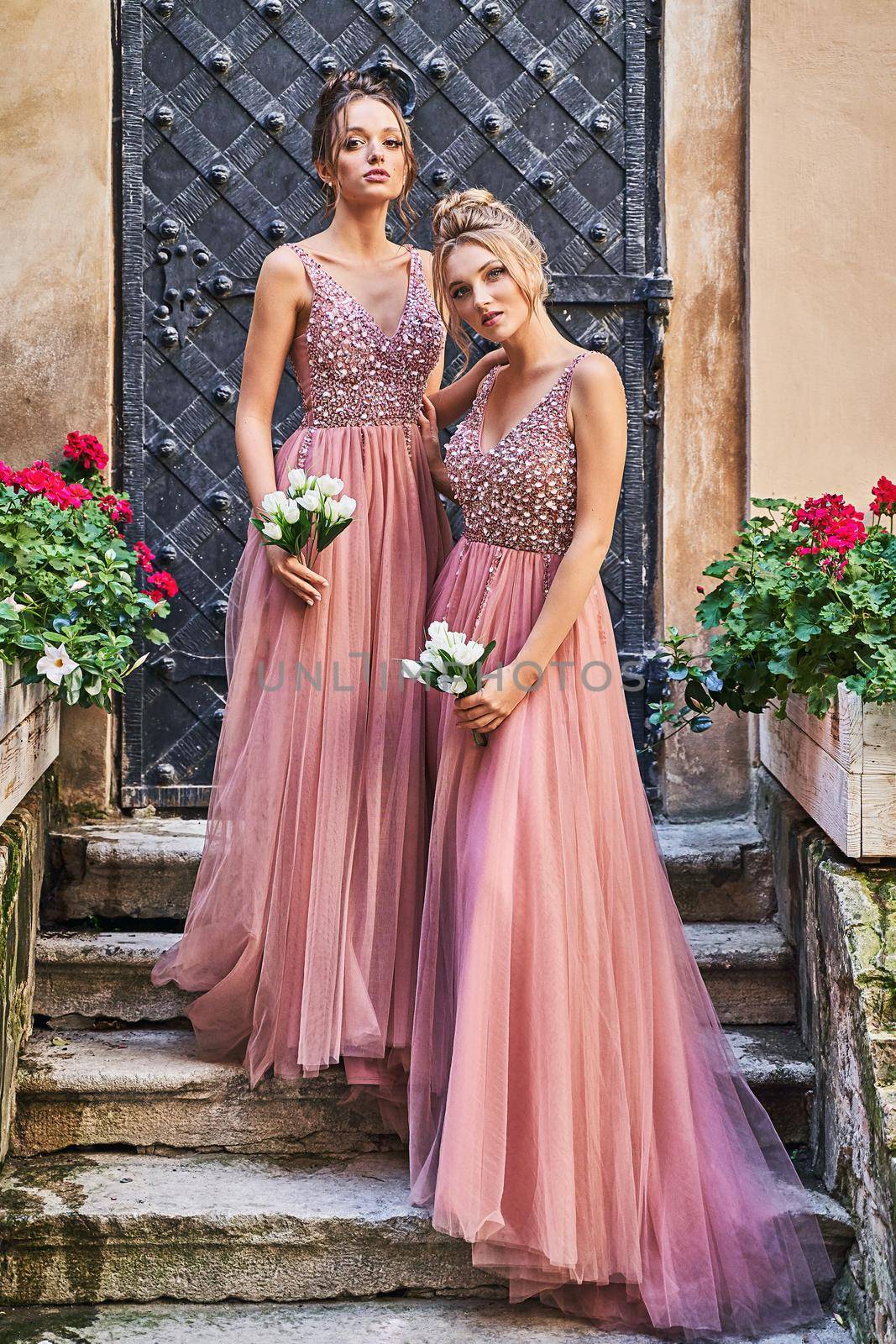 Beautiful bridesmaids in gorgeous elegant stylish red pink violet floor length v neck chiffon gown dress decorated with sequins sparkles and rhinestones holding flowers bouquets. Wedding day in old beautiful European city. by berezko