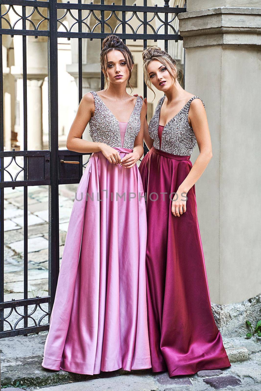 Two beautiful bridesmaids girls blonde and brunette ladies wearing elegant full length purple violet lilac lavender satin bridesmaid dress with silver sequined camisole top. European old town location for wedding day.