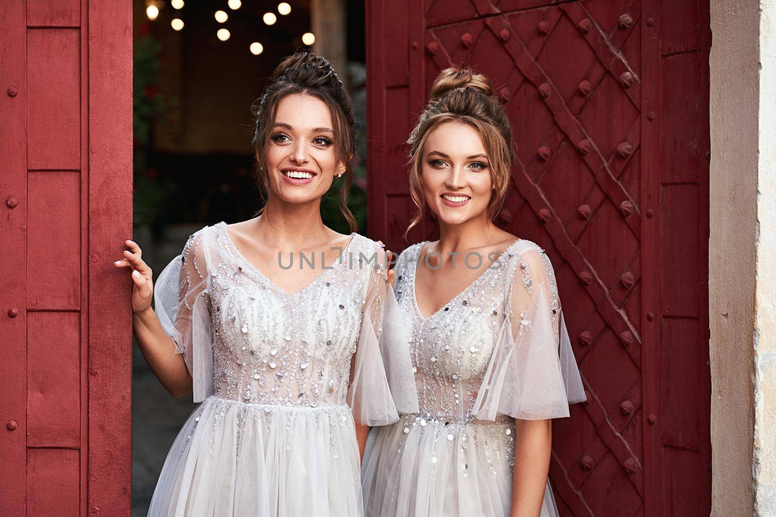 Beautiful retro styled bridesmaids ladies in gorgeous elegant stylish light grey silver floor length dresses in old beautiful European city welcome greeting guests at the gate. by berezko