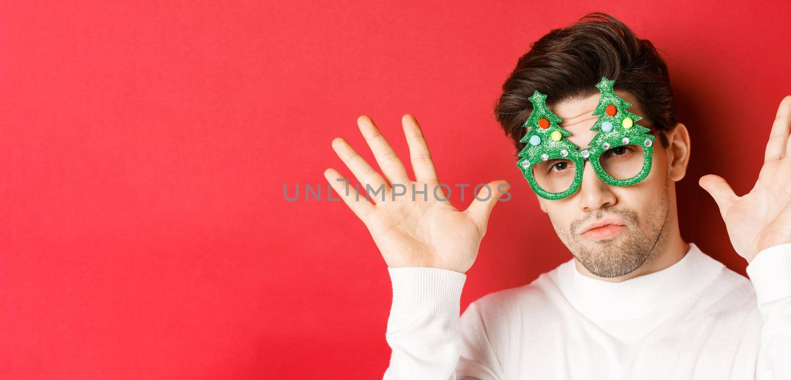 Concept of winter holidays, christmas and celebration. Close-up of funny handsome guy in party glasses, showing empty hands, standing over red background.