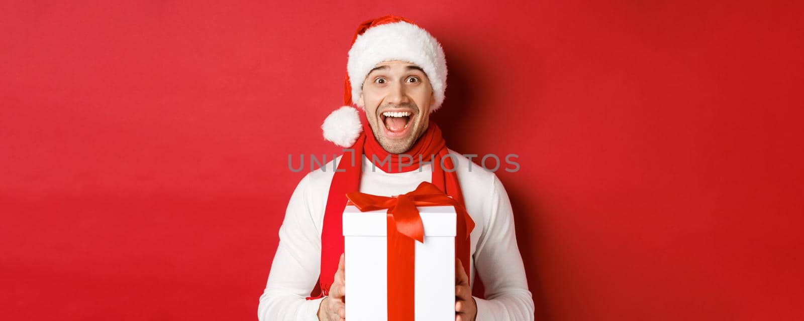 Concept of winter holidays, christmas and lifestyle. Close-up of excited handsome man in santa jat and scarf, looking amazed and receiving new year present, standing over red background by Benzoix