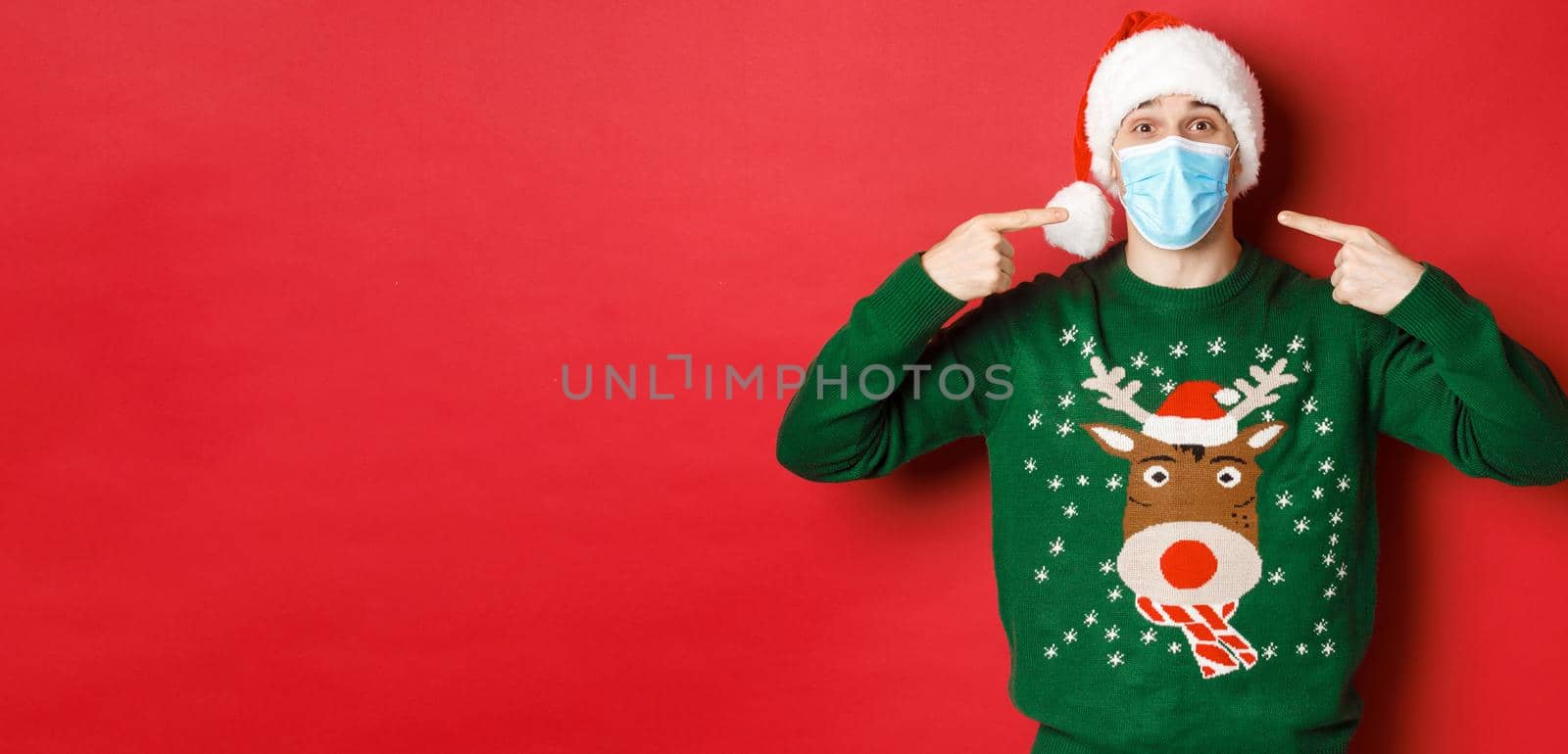 Concept of new year, coronavirus and social distancing. Happy man in santa hat and christmas sweater, recommending to wear medical mask on party, standing over red background.