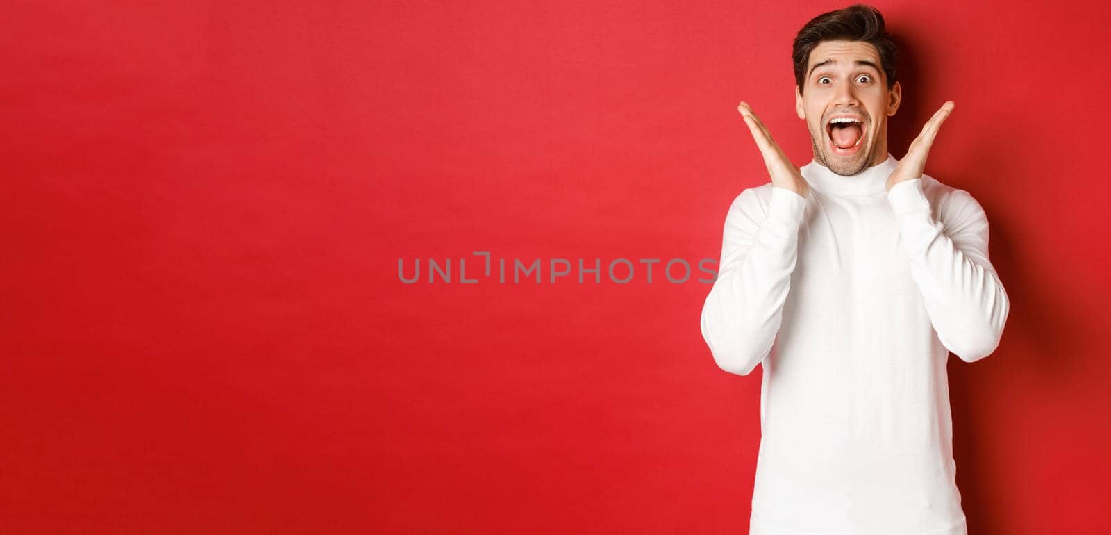 Concept of winter holidays, christmas and lifestyle. Surprised handsome man in white sweater, reacting to amazing new year promo offer, screaming amazed and raising hands, red background.