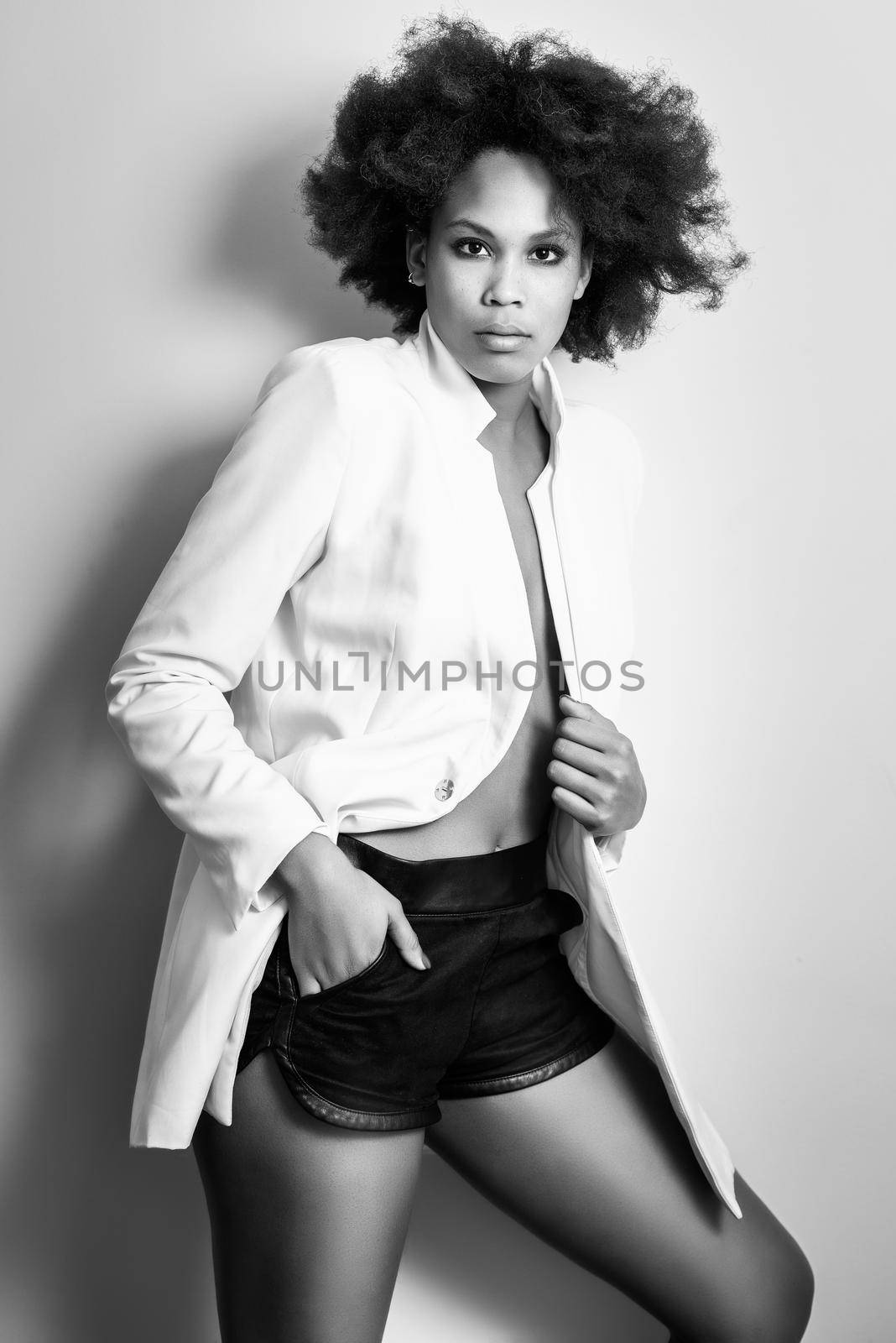 Young black woman with afro hairstyle possing near a white wall in her room. Mixed girl wearing blazer jacket and shorts.