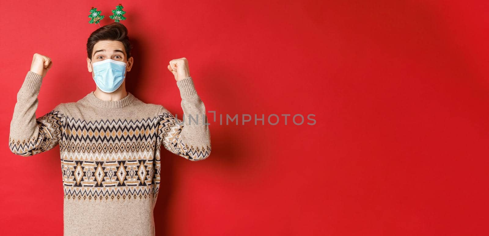 Concept of christmas, holidays and coronavirus. Happy man celebrating new year during covid-19 outbreak, wearing medical mask and sweater, rejoicing over red background by Benzoix