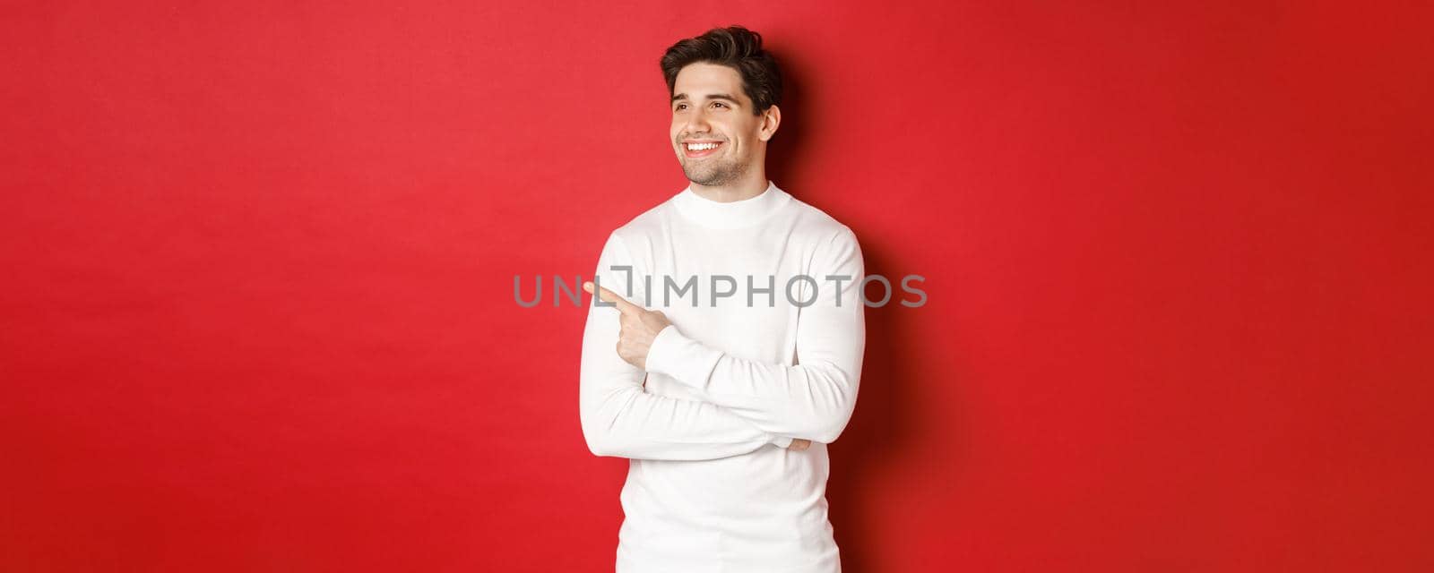 Concept of winter holidays, christmas and lifestyle. Handsome adult man in white sweater, looking and pointing left with pleased smile, showing new year advertisement, red background.