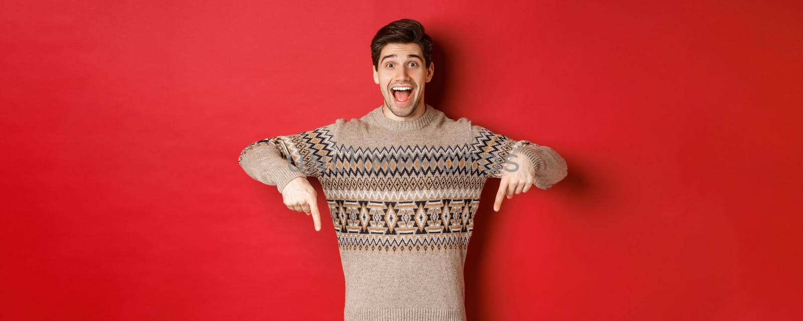 Concept of christmas celebration, winter holidays and lifestyle. Excited caucasian male model in xmas sweater, pointing fingers down and making announcement, showing advertisement, red background.