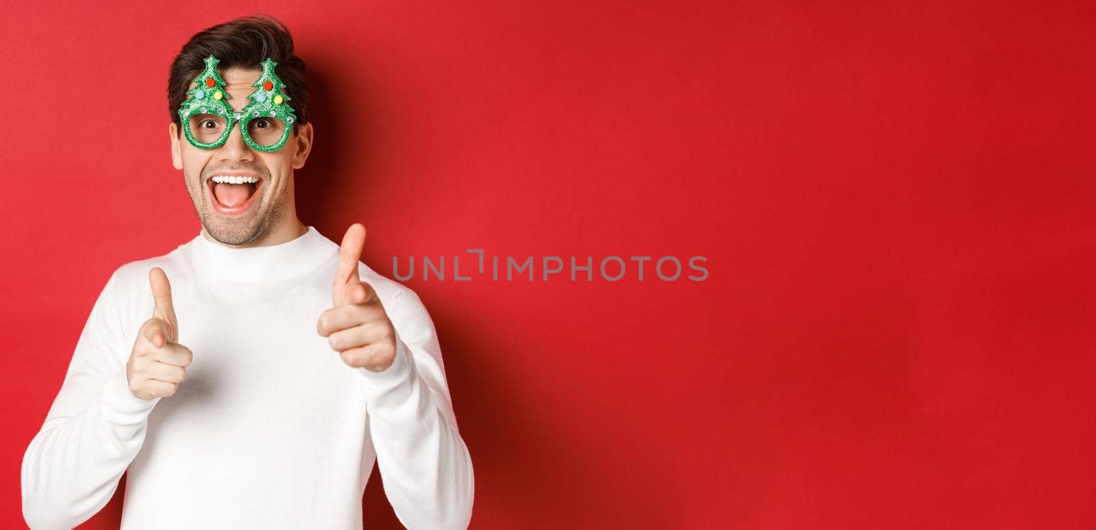 Joyful caucasian guy in party glasses and white sweater, smiling and pointing fingers at camera, wishing merry christmas and happy new year, standing over red background.