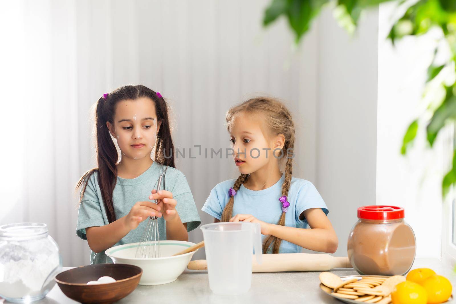 Two little girls in the kitchen prepare food, a dessert for the family. As they learn to cook they start playing with flour and smiling each other. Concept of: cooking classes, family, education.