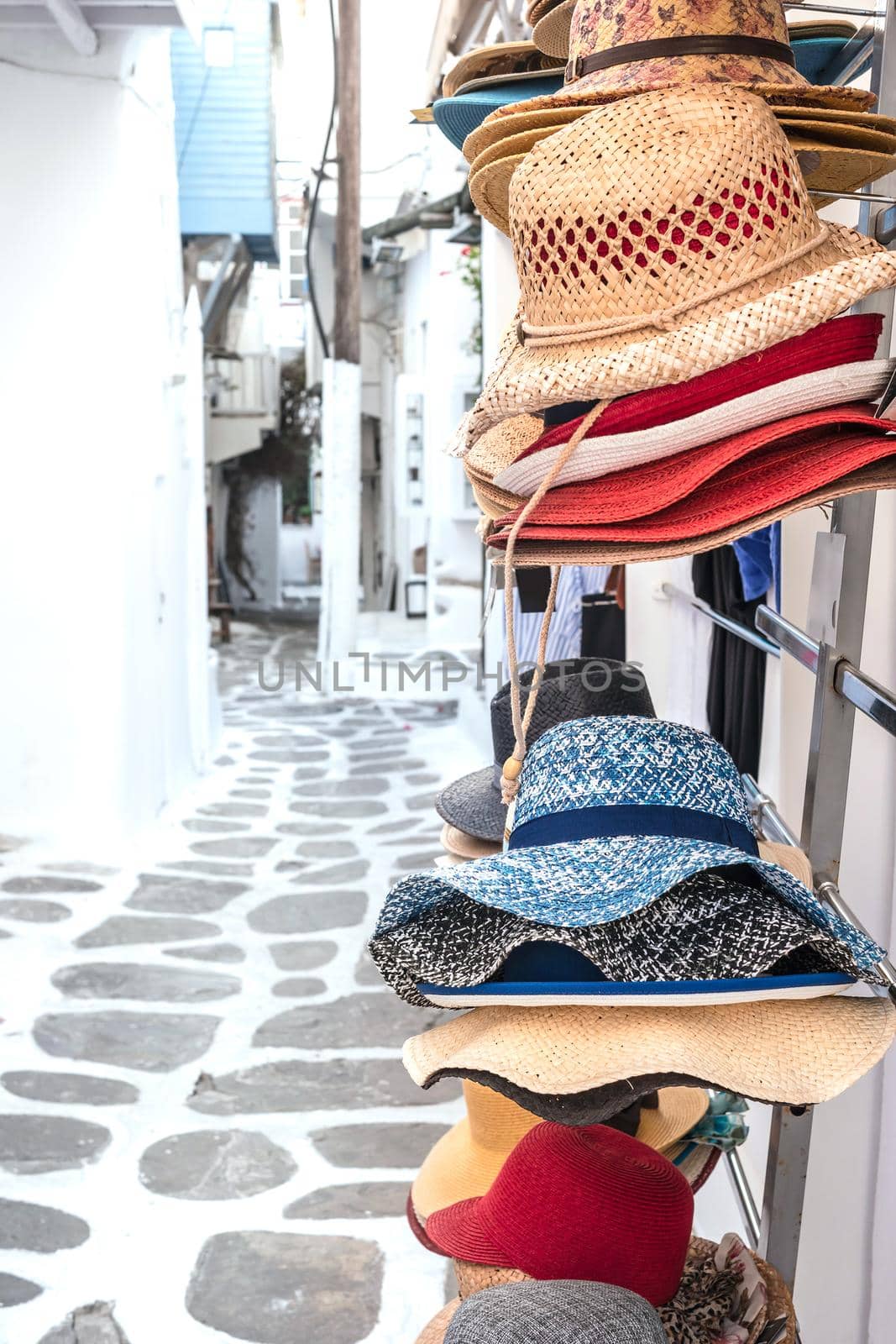 Colorful straw hats hanging on the market stall outdoor. Bright summer woman hats for sale on a street with white houses in Mikonas island, Greece.