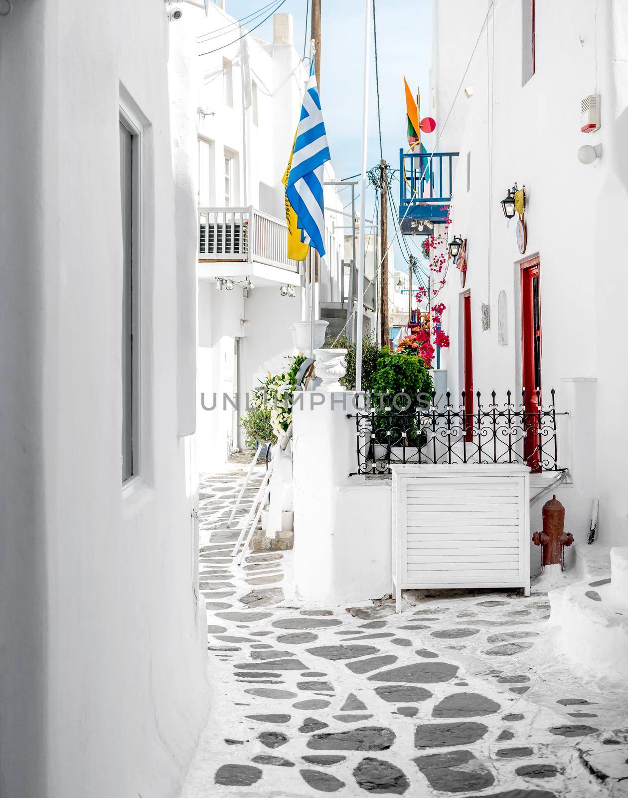 Neat narrow alley of old city on Mykonos island with traditional white houses and greek flag on pole
