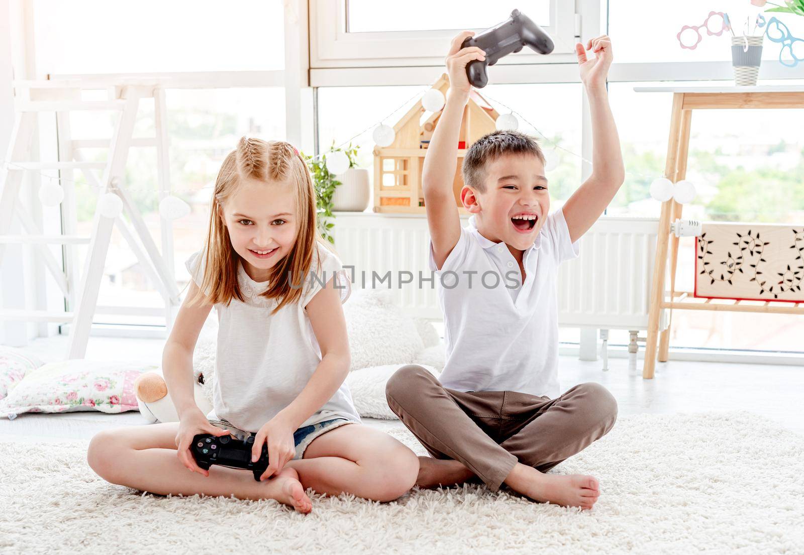 Cheerful little boy with hands up and cute little girl playing video game sitting in kids room