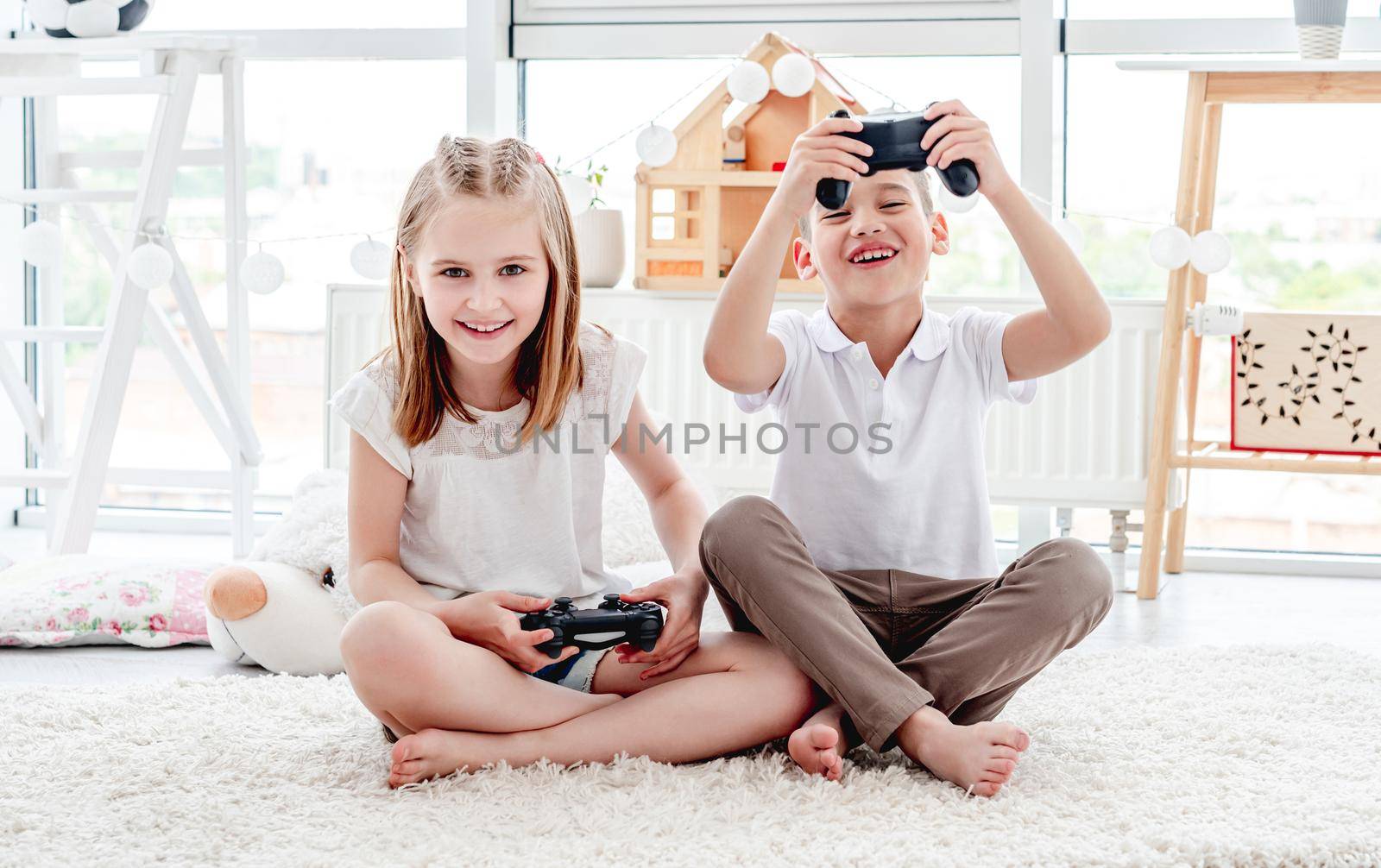 Playful kids with joysticks for video gaming sitting on floor in light room