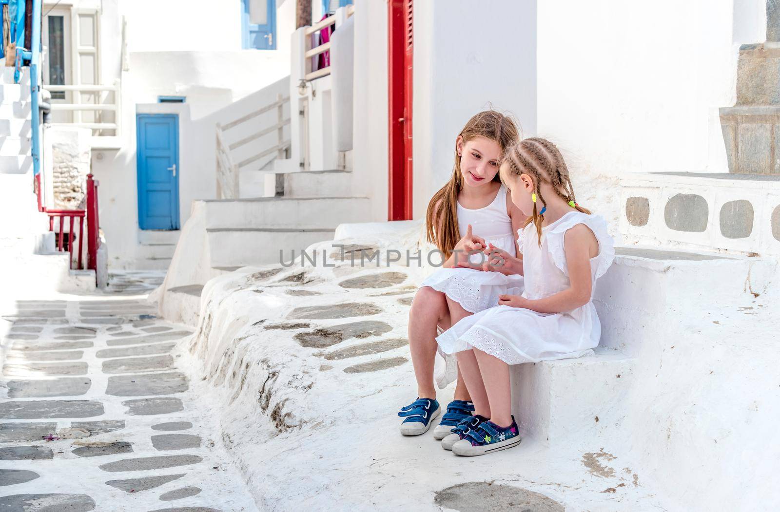 Two cute sisters dressed in white sitting on the stairs on street of greek traditional village with white houses