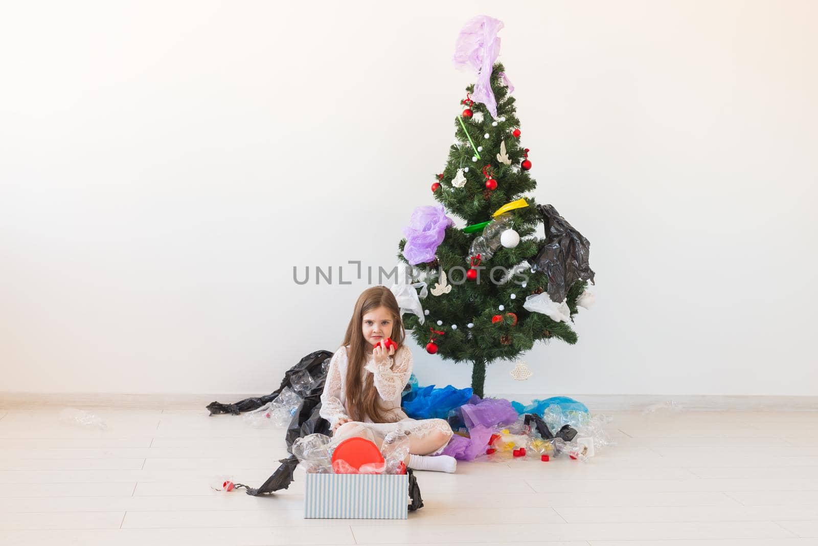 Shocked little child girl looks with opened eyes and worried expression, holding box with various plastic wastes over christmas tree background. by Satura86