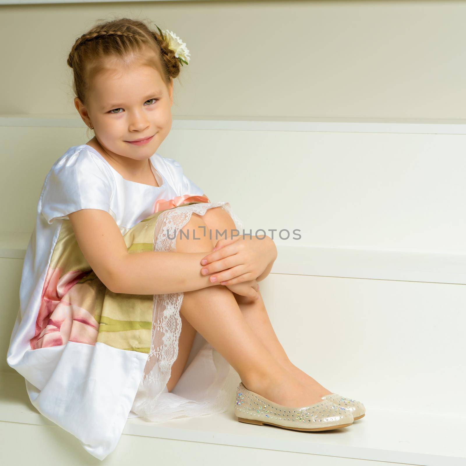 A charming little girl in a smart dress sits on a white staircase. The concept of happiness, childhood.