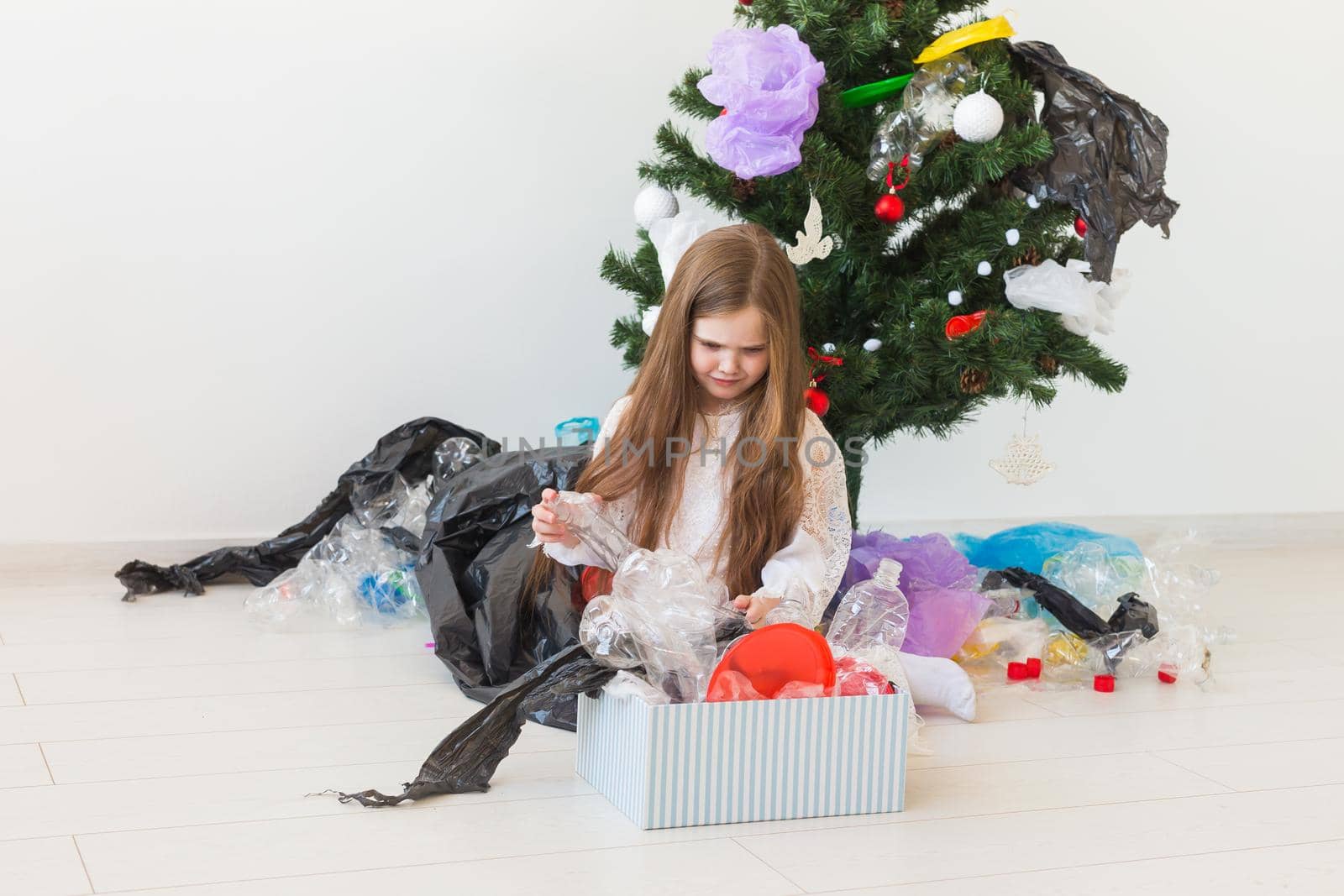 Shocked little child girl looks with opened eyes and worried expression, holding box with various plastic wastes over christmas tree background