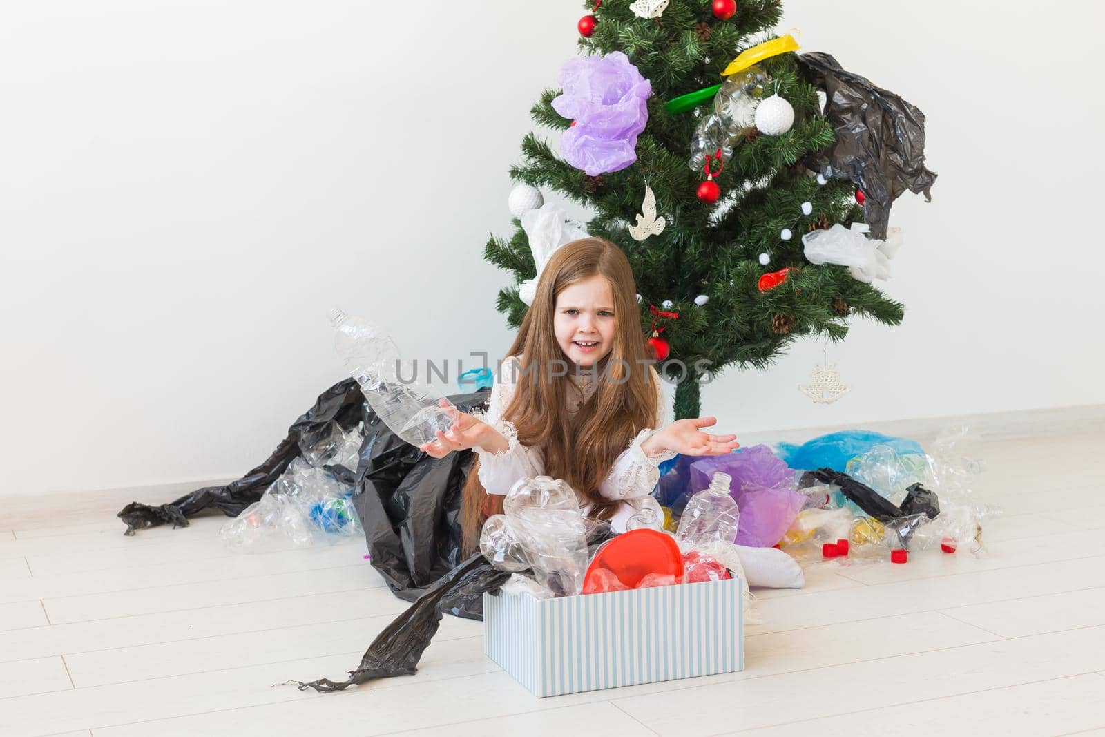 Shocked little child girl looks with opened eyes and worried expression, holding box with various plastic wastes over christmas tree background. by Satura86
