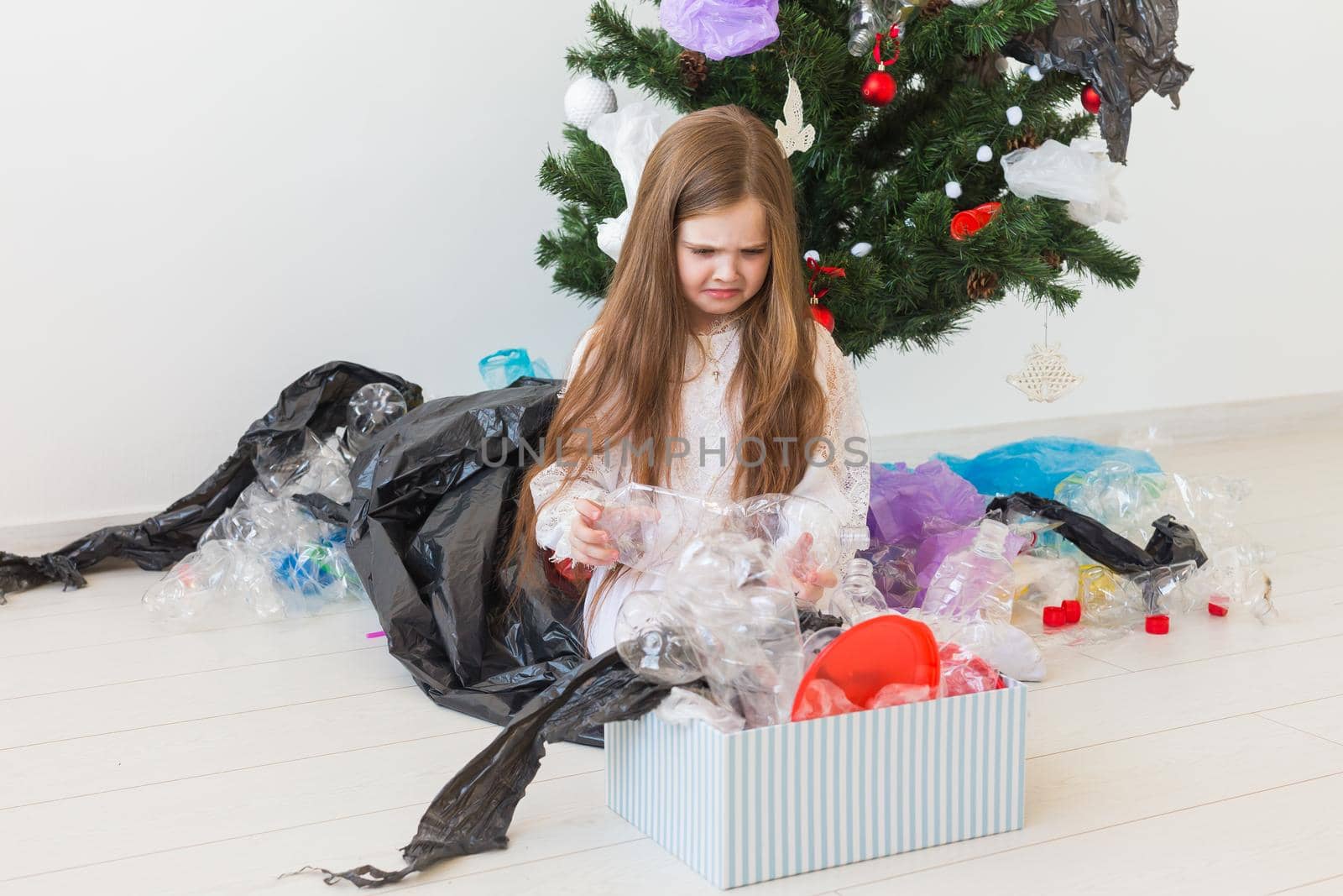 Shocked little child girl looks with opened eyes and worried expression, holding box with various plastic wastes over christmas tree background